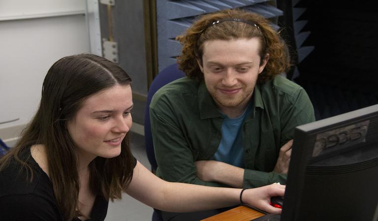 学生 sit side by side smiling and looking at a computer screen.